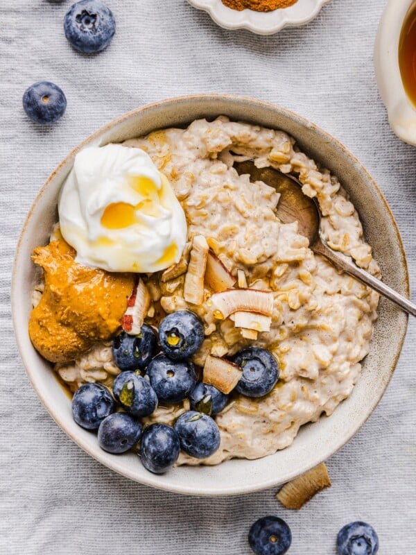 White bowl with greek yogurt oatmeal, topped with blueberries, nut butter, and extra yogurt.