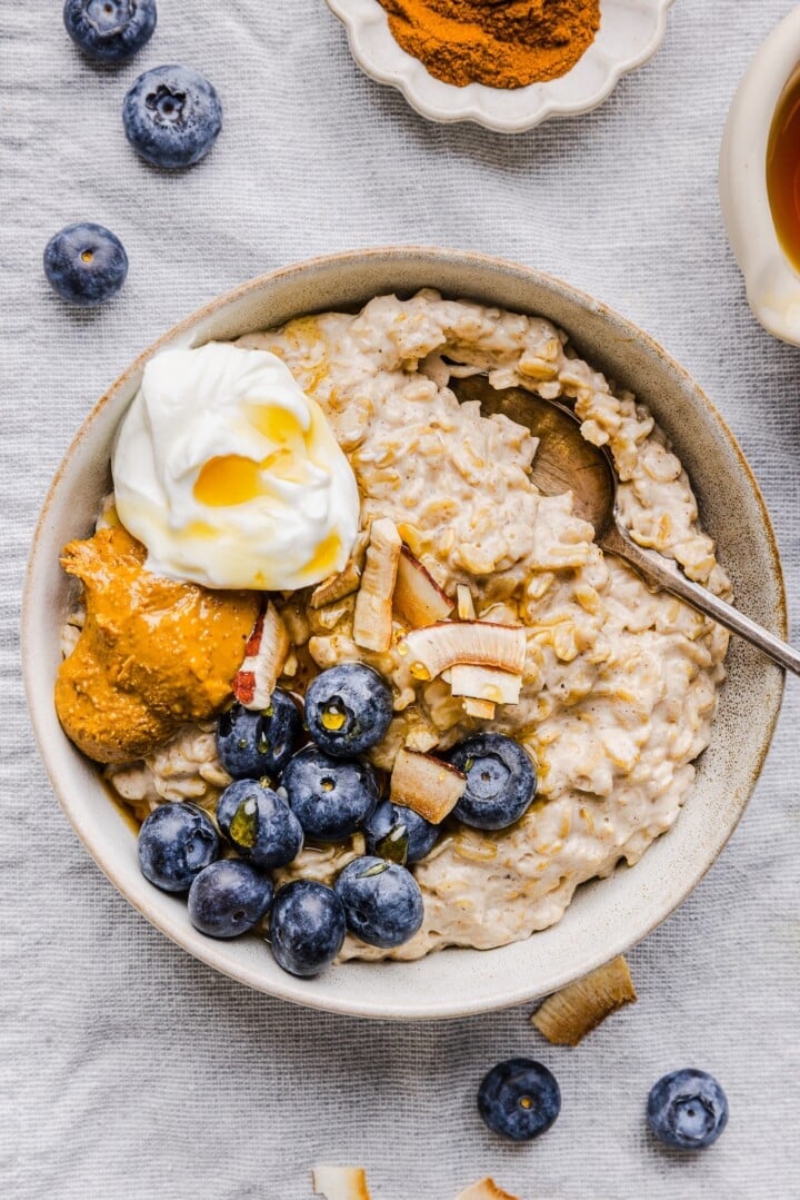 White bowl with greek yogurt oatmeal, topped with blueberries, nut butter, and extra yogurt.