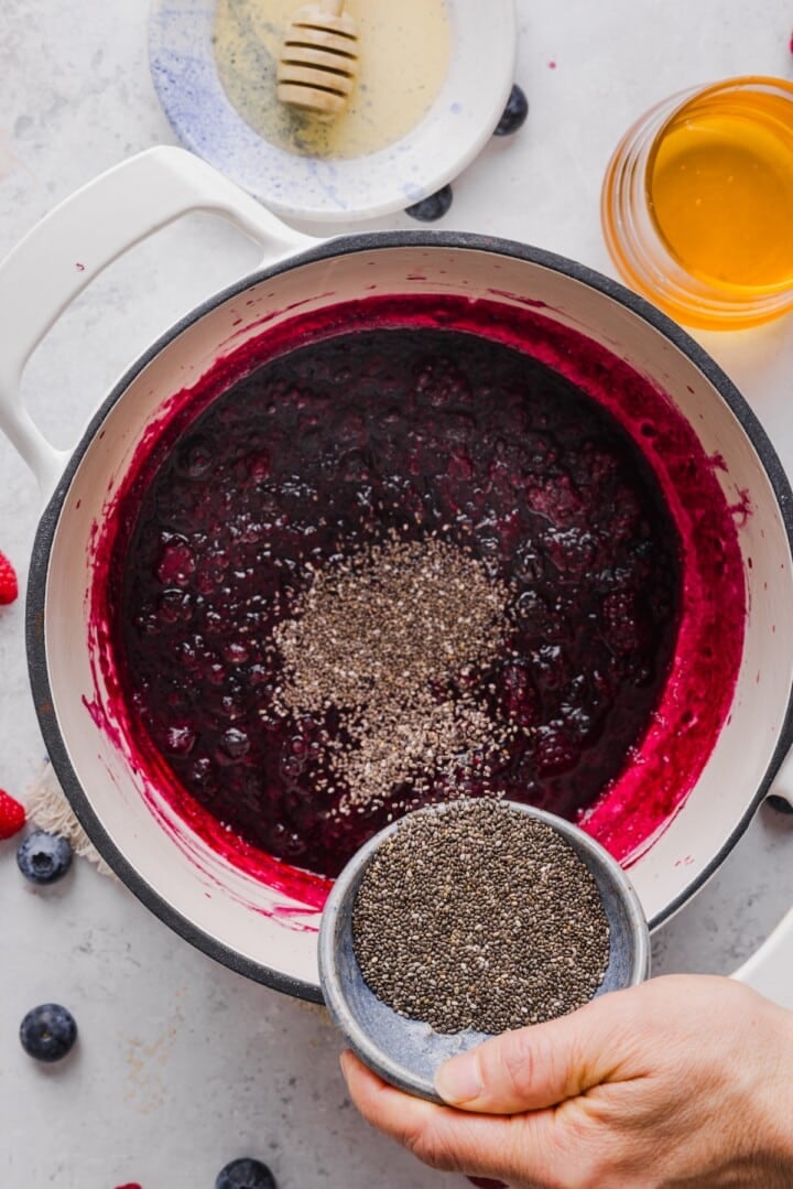 Frozen berries melted in a pan on the stove, with a woman's hands pouring chia seeds in.