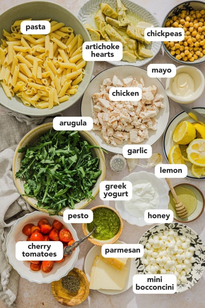 Ingredients for pesto chicken pasta salad displayed in small bowls on a marble counter.