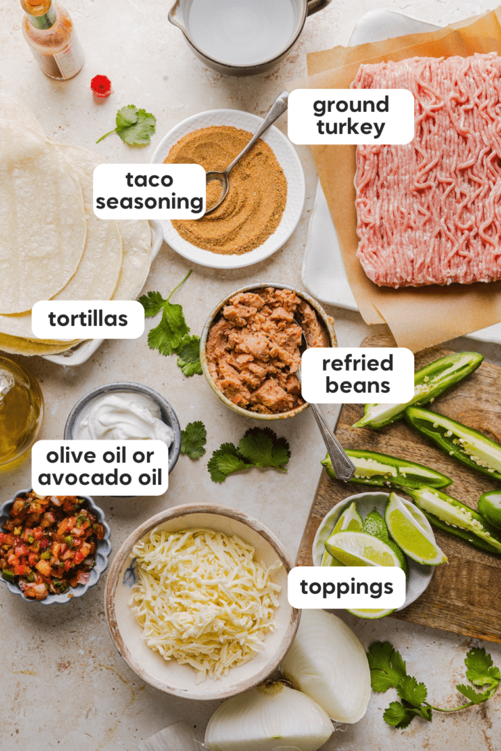 Ingredients for turkey tacos including ground turkey, refried beans, tortillas, shredded cheese, olive oil, taco seasoning, jalapeños, lime, crema, and cilantro.