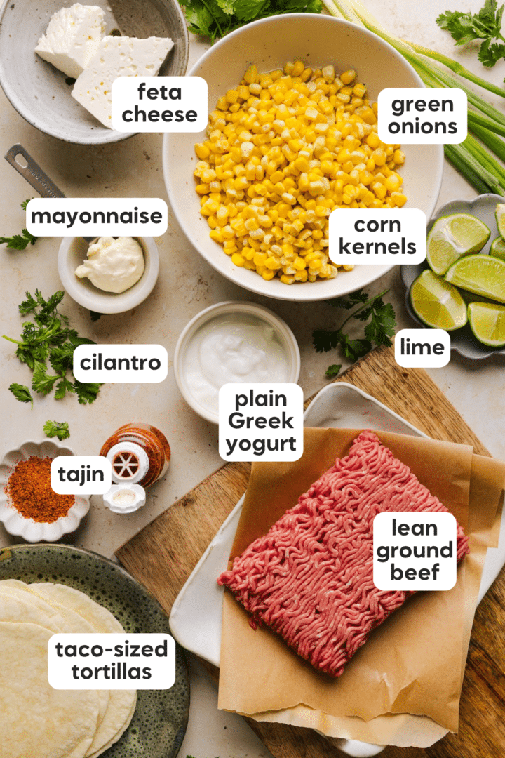 Ingredients for street corn tacos including corn, lean ground beef, plain Greek yogurt, mayonnaise, lime juice, tajin, green onions, feta cheese, and taco-sized tortillas.